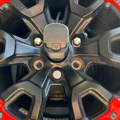 LensJet OEM nozzle housing installed on a Ford Bronco rear camera, viewed straight on through a black and red Bronco Raptor spare tire wheel with the washer tubing routed through the spokes.