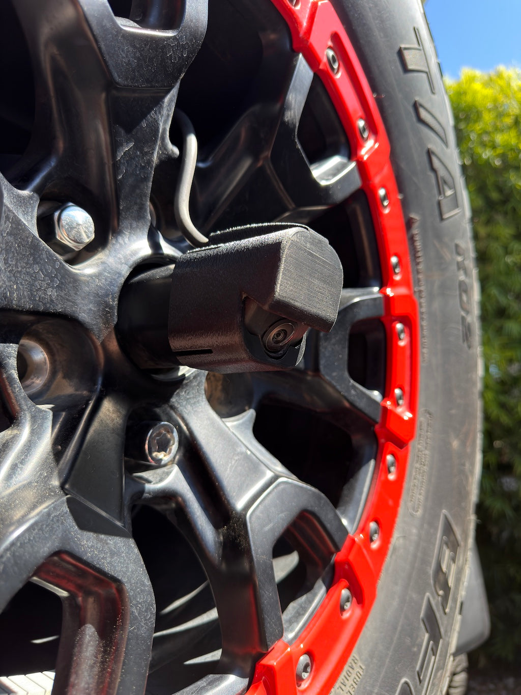 Close-up side view of the LensJet OEM nozzle housing installed on a Ford Bronco rear camera, showing the housing profile and camera lens through a black and red Bronco Raptor spare tire wheel.