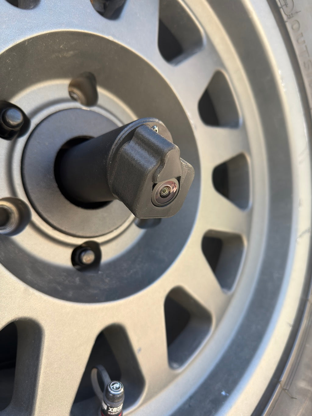 Side view of the LensJet Turn Offroad nozzle housing installed on a Ford Bronco rear camera stalk, showing the housing profile and camera lens through an aftermarket spare tire wheel spokes.