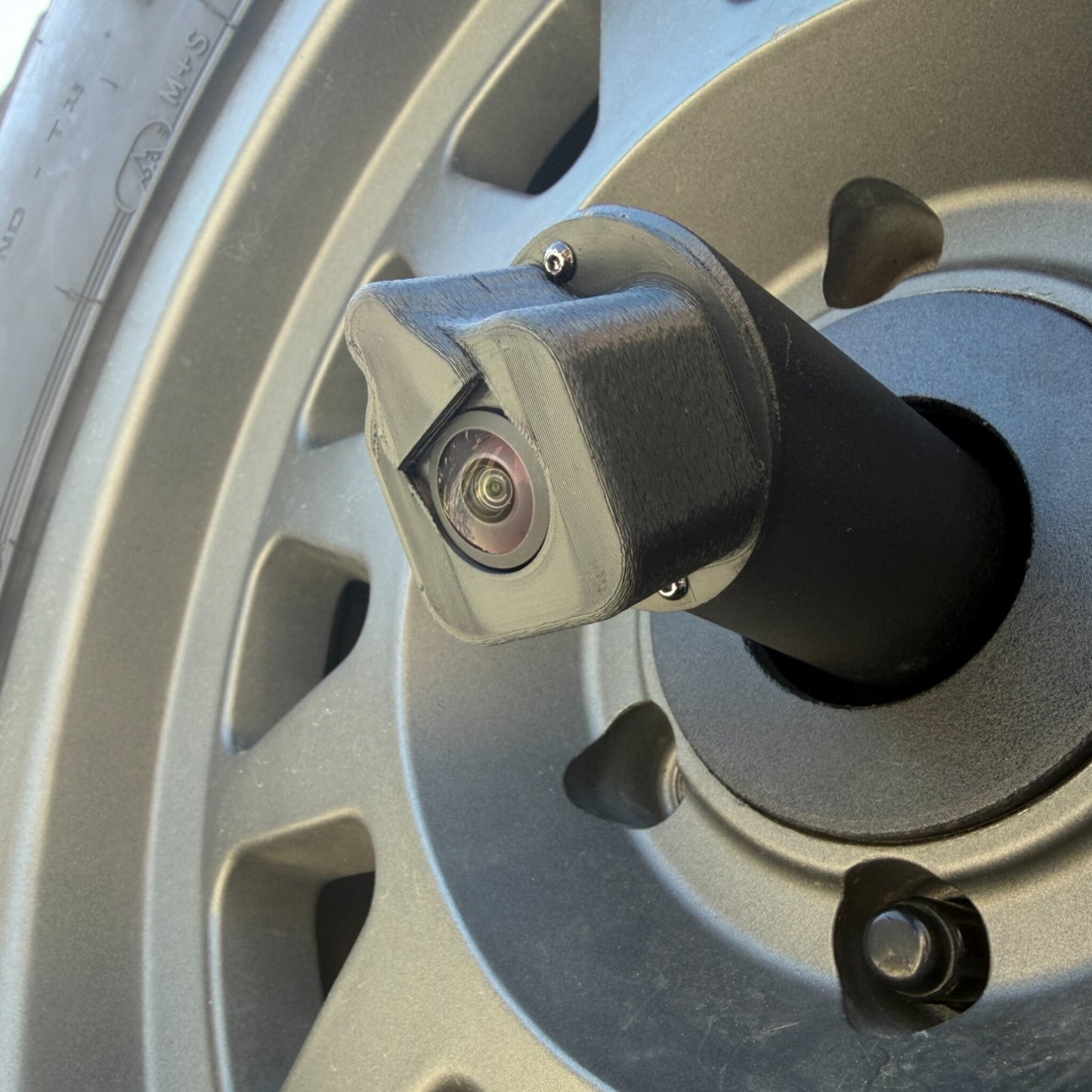 LensJet Turn Offroad nozzle housing installed on a Ford Bronco rear camera stalk, viewed through the spare tire wheel spokes. The housing fits over the camera lens with a built-in spray nozzle opening.