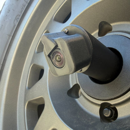 LensJet Turn Offroad nozzle housing installed on a Ford Bronco rear camera stalk, viewed through the spare tire wheel spokes. The housing fits over the camera lens with a built-in spray nozzle opening.
