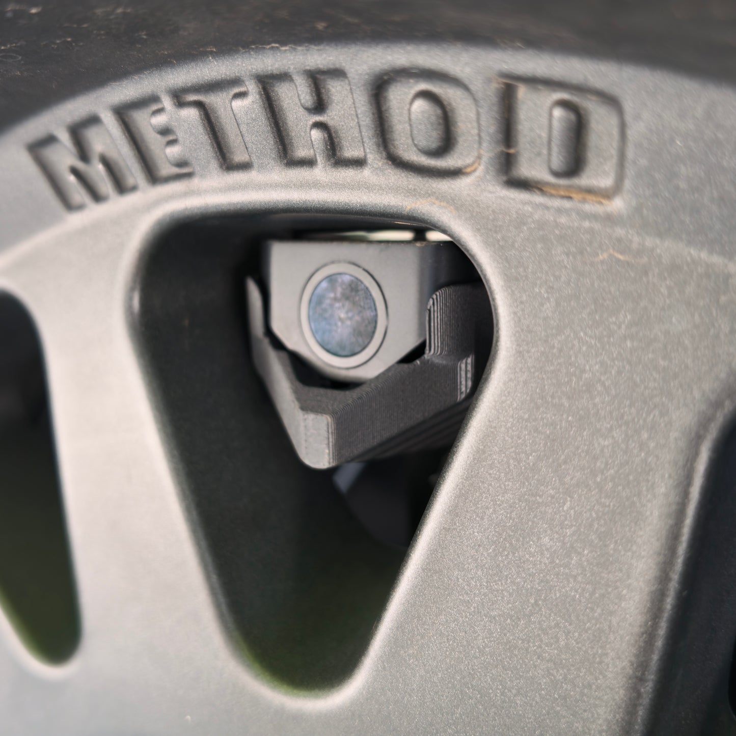 Close-up of the LensJet for Wolfbox mounting bracket and hardware framed by a Method wheel spoke